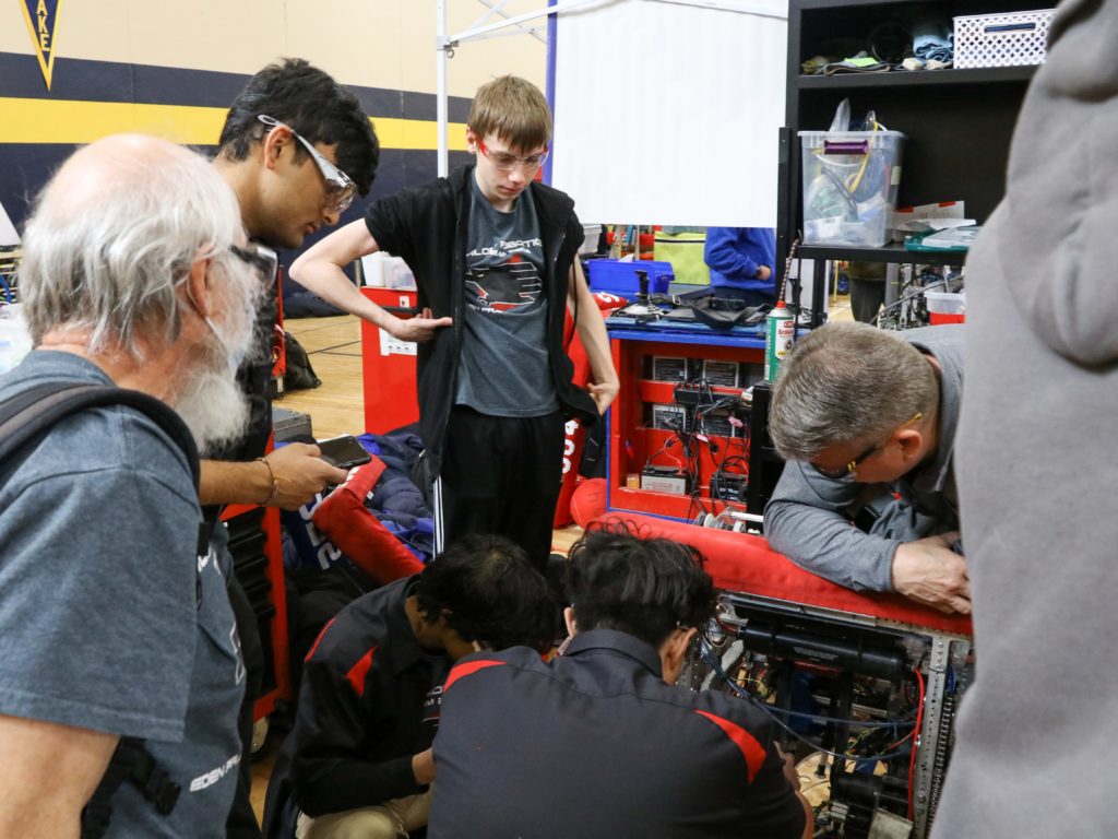 Multiple mentors and students huddling around the robot as two students attempt to fix the robot. 