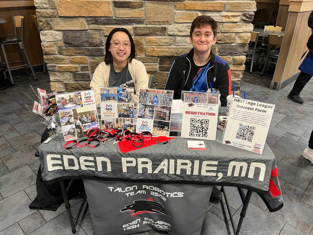 Two team members sitting down next to a table and smiling. the table has various items that lists off information about FIRST. The table also has a banner that says Talon Robotics.
