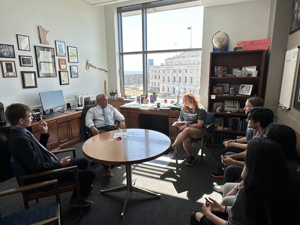 7 people talking in a room, there is a window that is shining sunlight inwards. everyone is smiling and in mid-conversation. 