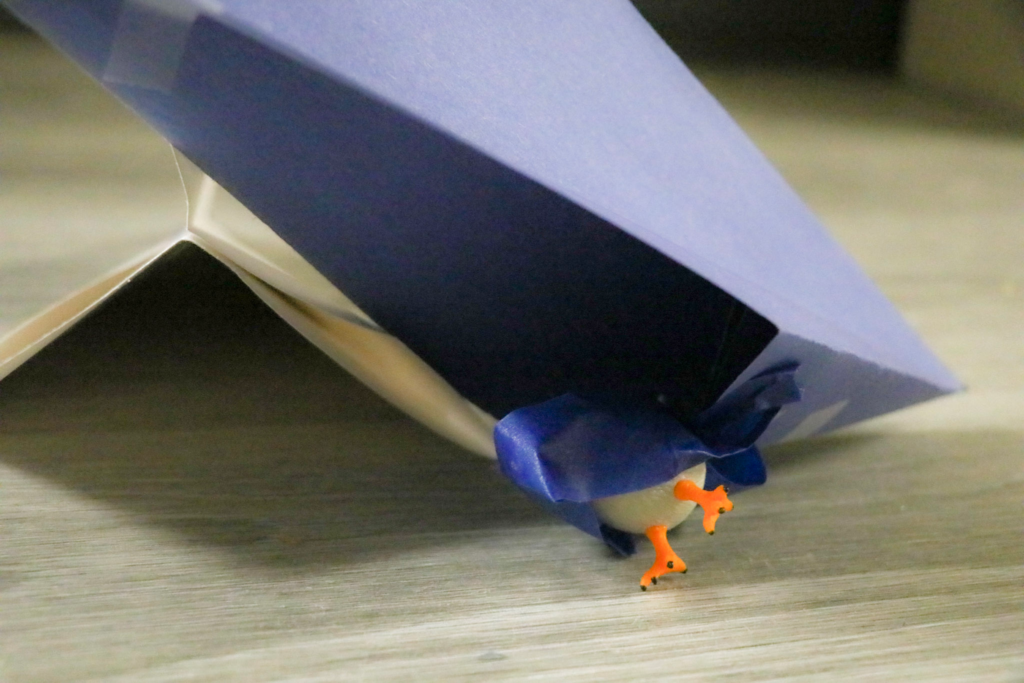 A plastic pigeon poorly taped to a paper airplane. The plane has hit the ground.