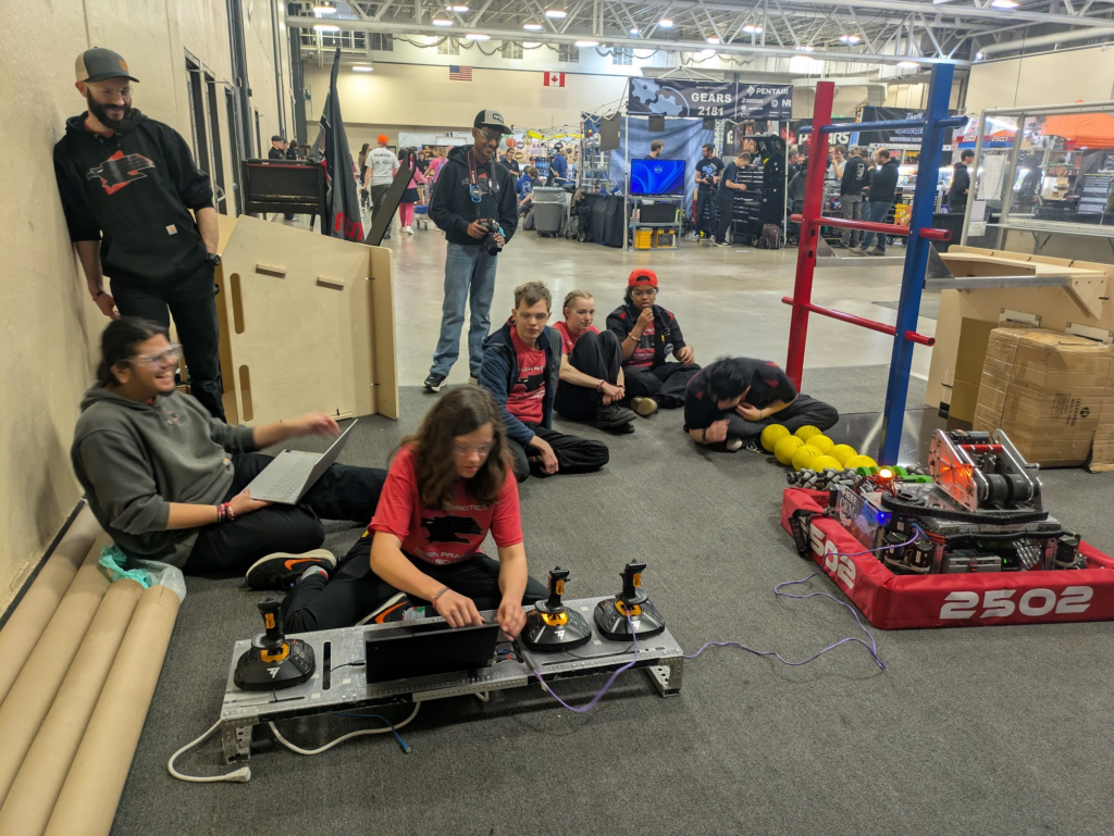 Drive team and mentors sitting on the practice field as a programmer works on the bot