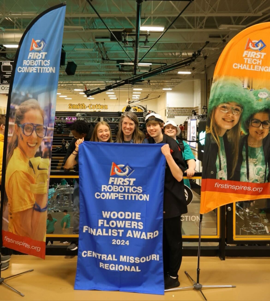 Ally, Lisa, and Riley posing with Lisa's Woodie Flowers blue banner. Riley's friend Luke can be seen posing in the background